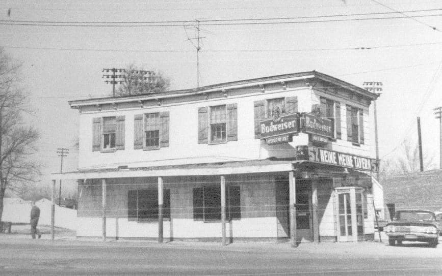 Black and white photograph of Heine Meine Tavern, a two-story frame building with Budweiser signs and a sedan parked at the curb
