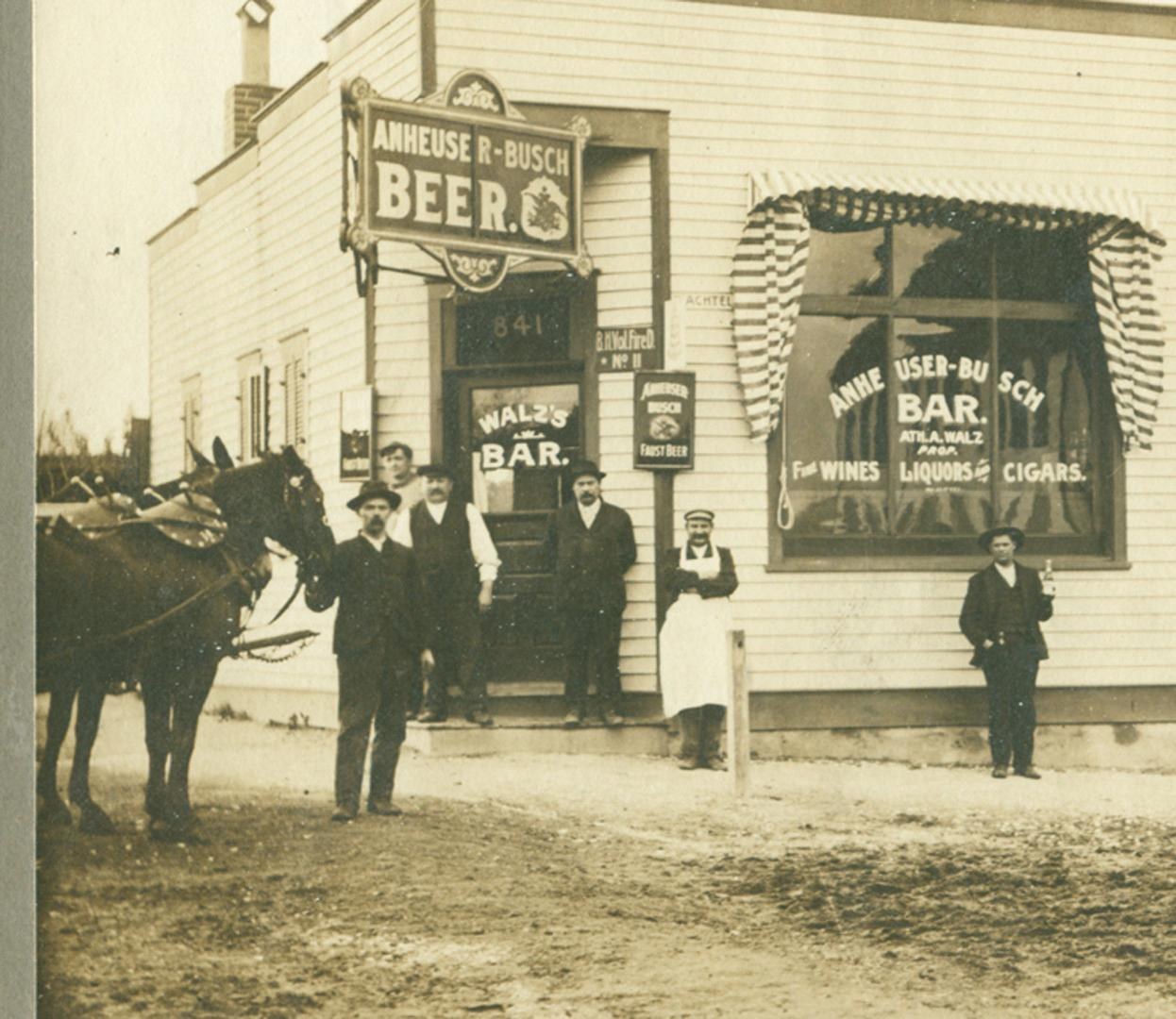 Sepia photograph of Walz's Bar, a white frame saloon with an Anheuser-Busch Beer sign over the door, six men and a team of horses posed out front on a dirt street