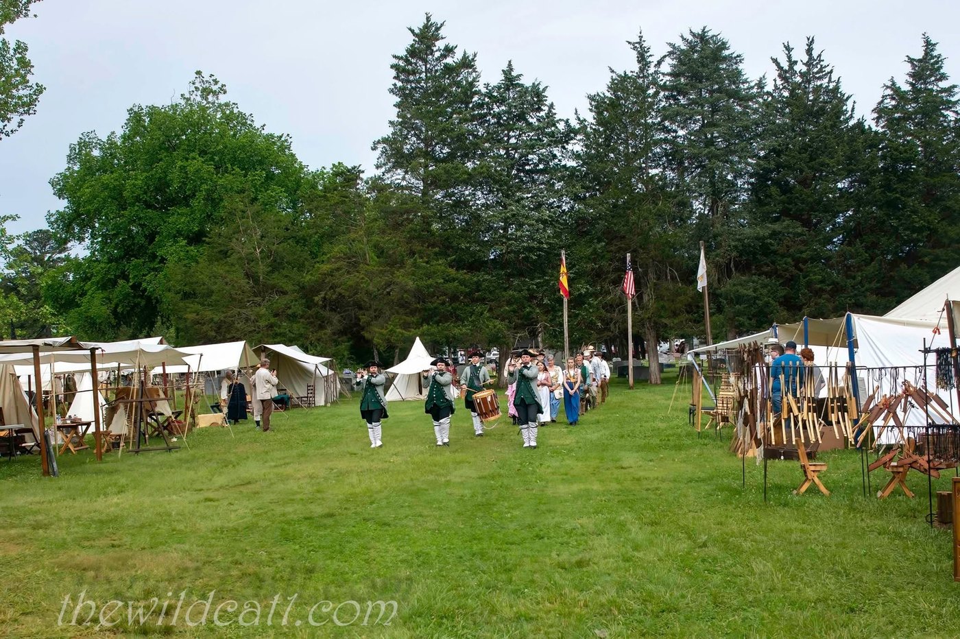 Reenactors in green coats and tricorn hats march in a line across a grass field toward white canvas tents and flags