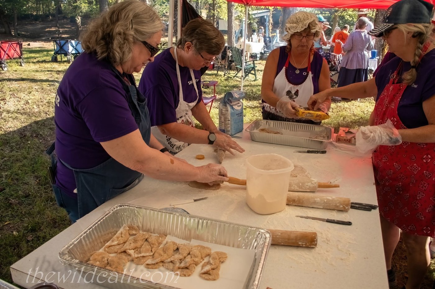 Volunteers in purple shirts and aprons roll and cut dough at a long table under a red canopy, with a tray of finished pastries in front