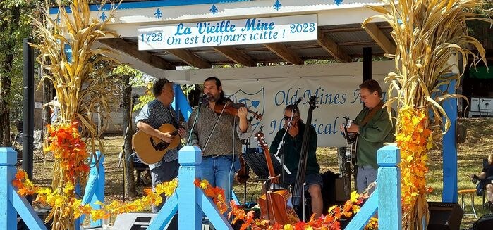 A fiddler, guitarist, bassist and banjo player on a blue bandstand decorated with corn stalks and fall leaves, under a banner reading La Vieille Mine 1723-2023, On est toujours icitte!
