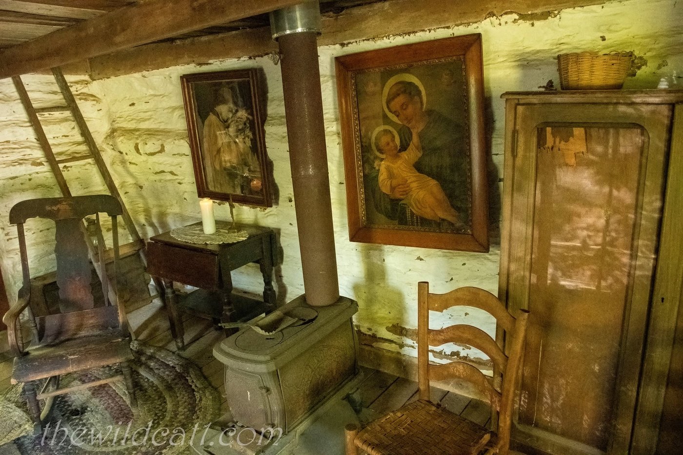 Inside a log cabin: a wood stove, ladder-back chairs, a braided rug and framed religious pictures on the log walls