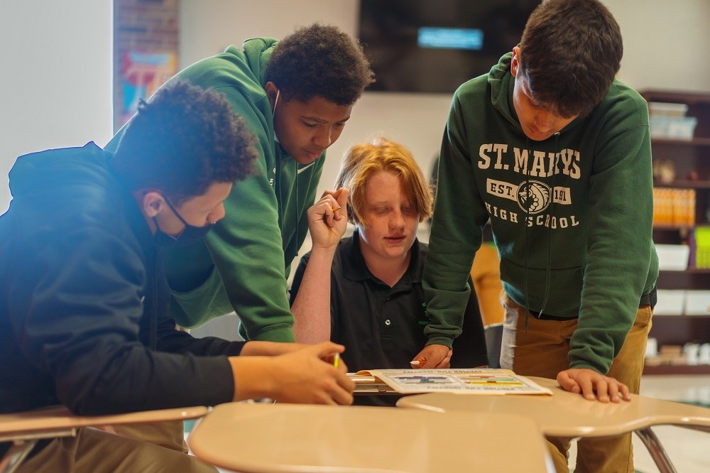 Four students in green St. Mary's hoodies leaning over a desk working on an assignment together