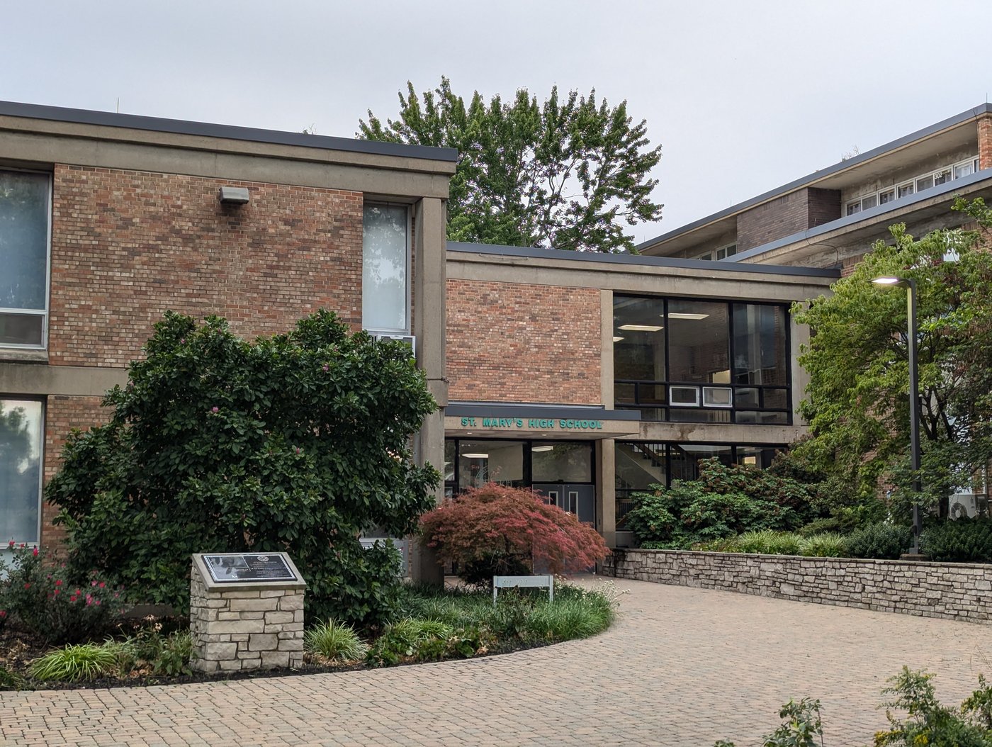 Two-story brick school building with the words ST. MARY'S HIGH SCHOOL above a glass entrance, landscaping and a stone memorial out front