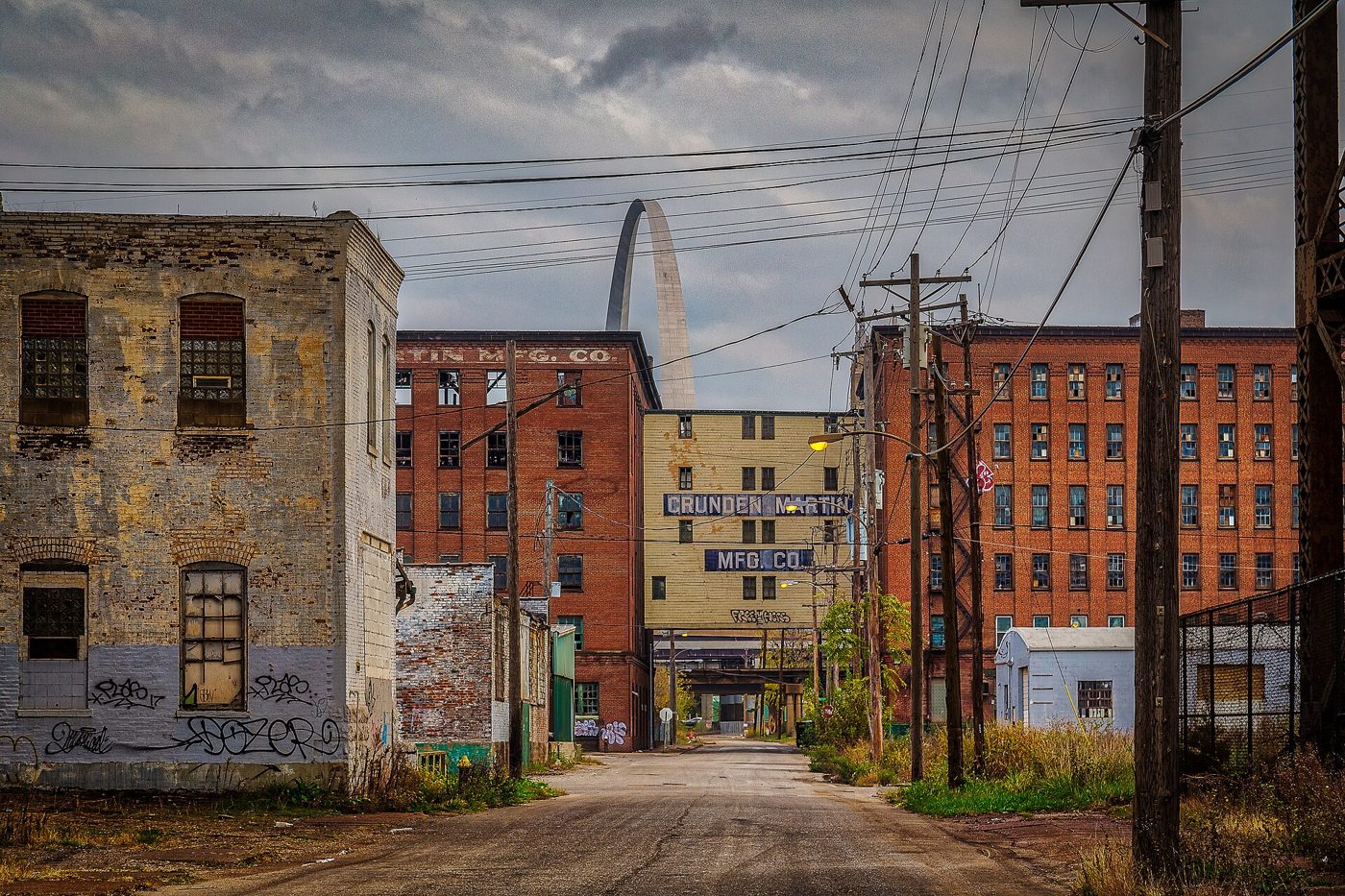 A gritty city street lined with old brick warehouses and utility poles, with the Gateway Arch rising in the distance under a cloudy sky