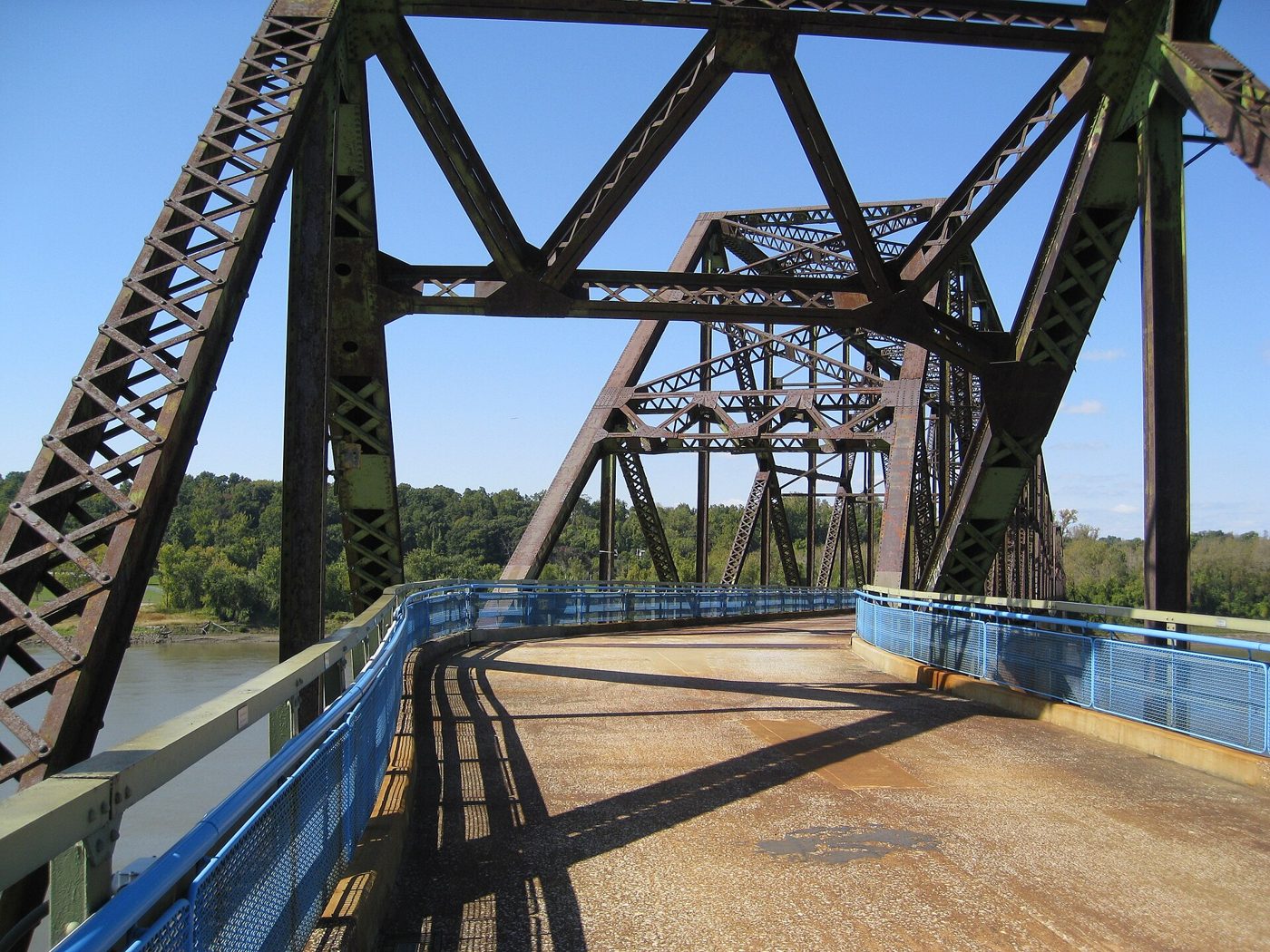 Looking along the deck of a steel truss bridge painted gray-green, with blue railings, where the deck visibly turns to the left partway across the river