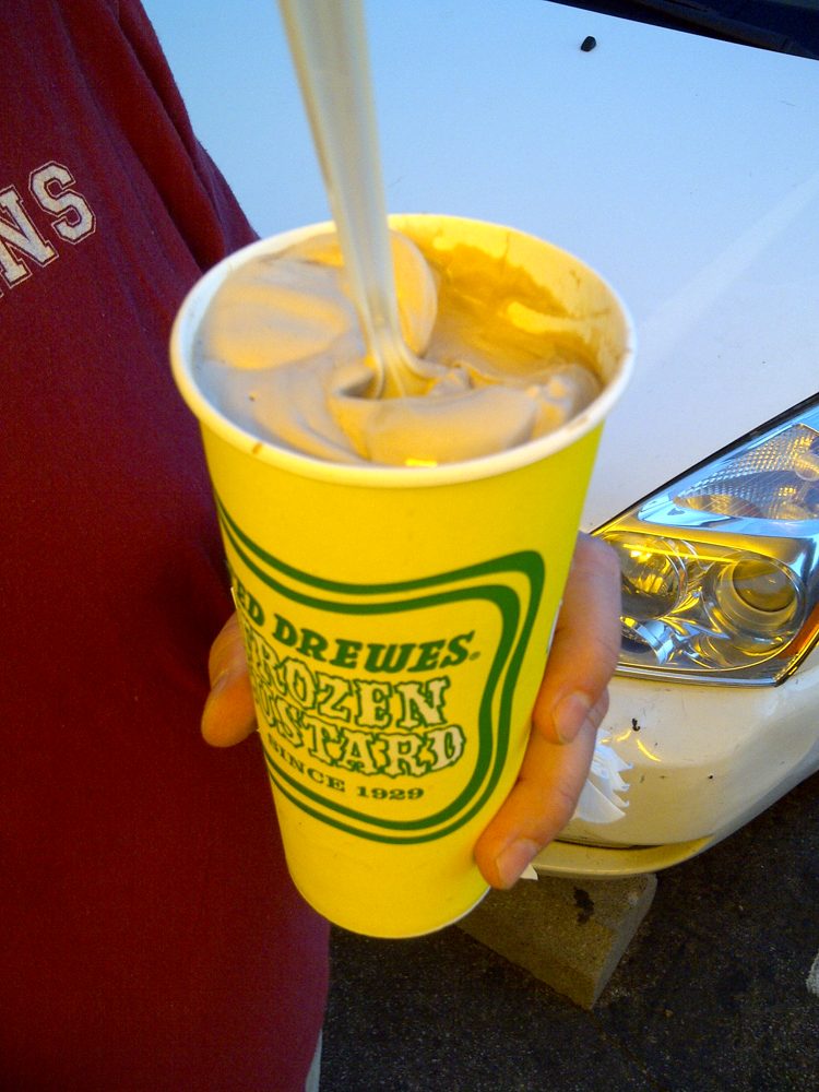 A hand holding a yellow Ted Drewes cup of frozen custard with a spoon standing straight up in it