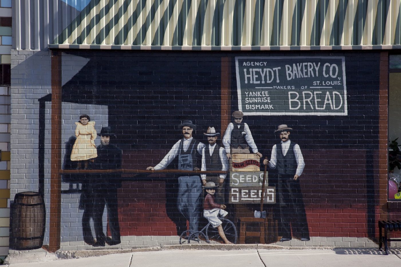 A painted mural of five men in 1900s work clothes and a boy with a bicycle standing in front of a brick storefront under a Heydt Bakery Co. Bread sign
