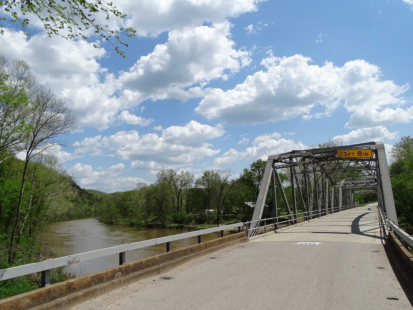 A narrow steel truss bridge with a curved approach, painted silver-gray, crossing a green river with wooded bluffs behind it
