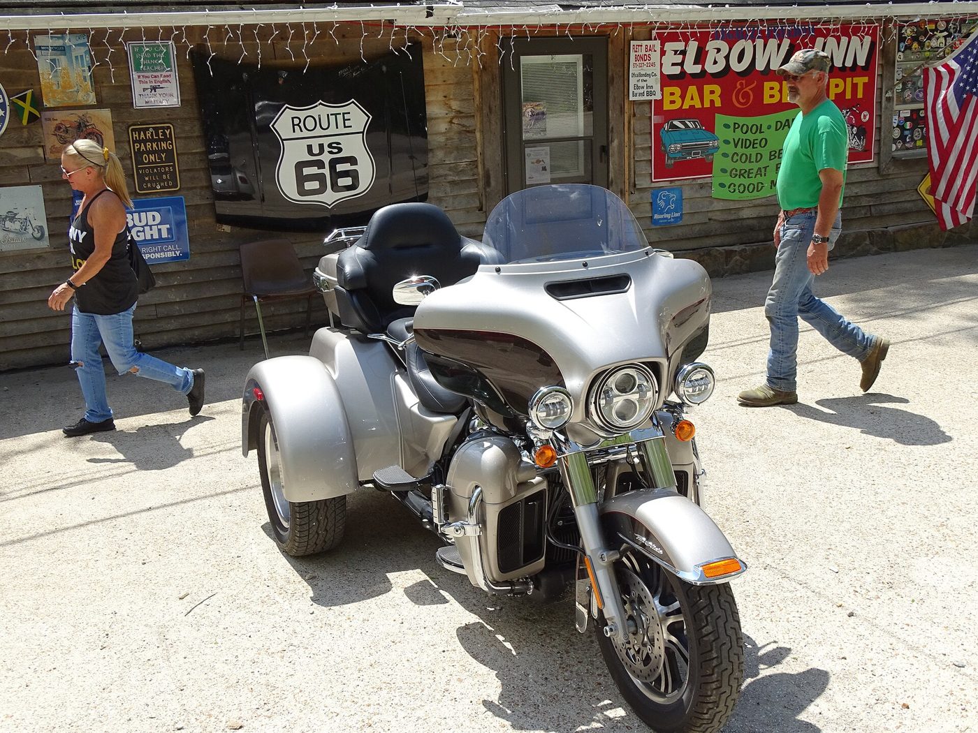 A low stone roadhouse with an Elbow Inn Bar and BBQ Pit sign and a Route 66 shield, a silver trike motorcycle and two people out front