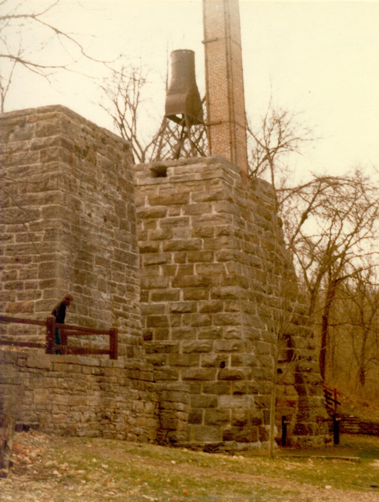 A tall square stone furnace stack with a small iron door at its base, standing among bare trees