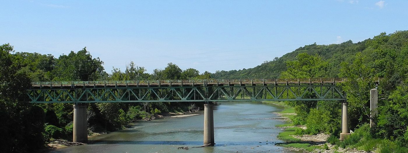 A long steel truss bridge on concrete piers spanning a wide brown river, with wooded hills behind