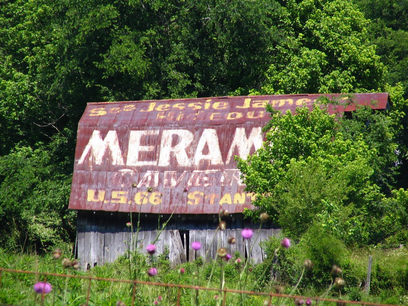 An old weathered barn with a faded red roof painted with the words Jesse James Hideout, Meramec Caverns, U.S. 66 Stanton