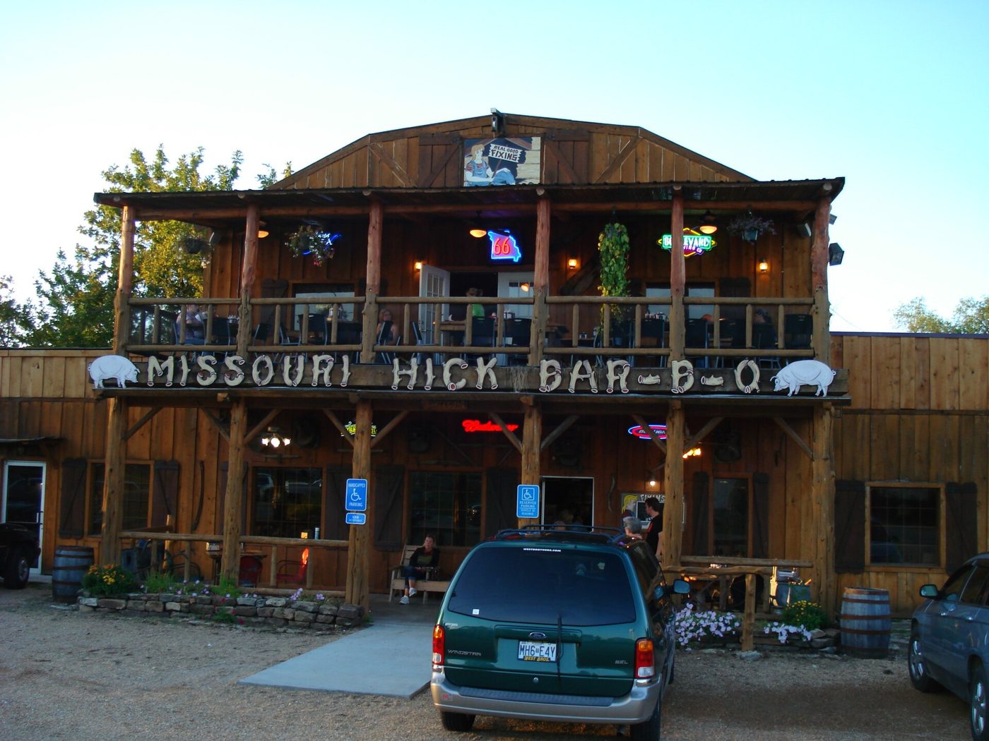 A two-story log restaurant with a wooden sign reading Missouri Hick Bar-B-Q, a covered porch with rocking chairs, and a green minivan parked in front at dusk