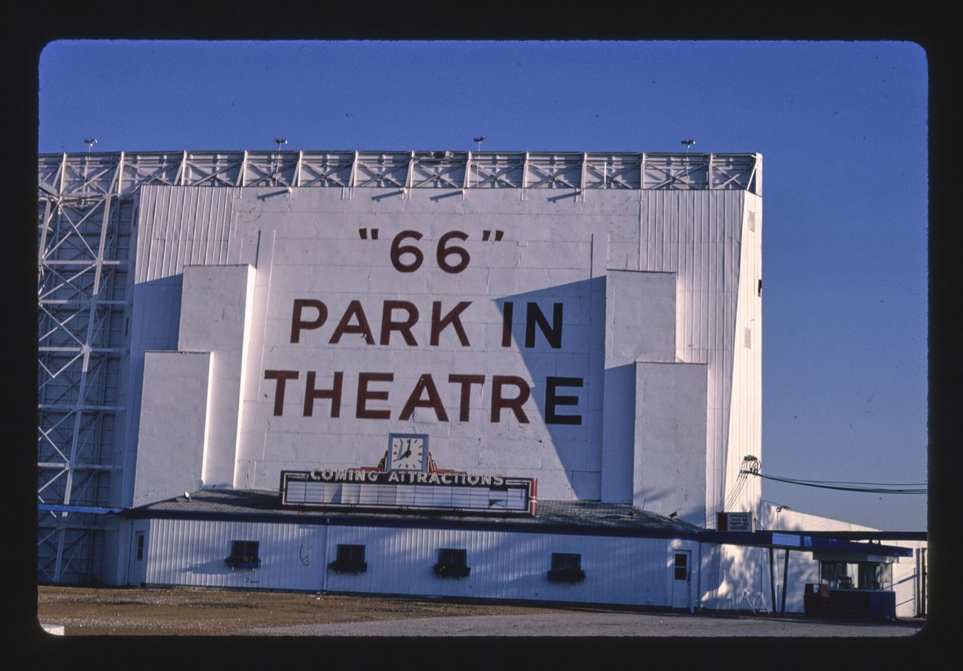 A tall white drive-in theater screen tower with the words 66 Park In Theatre in black letters against a deep blue sky