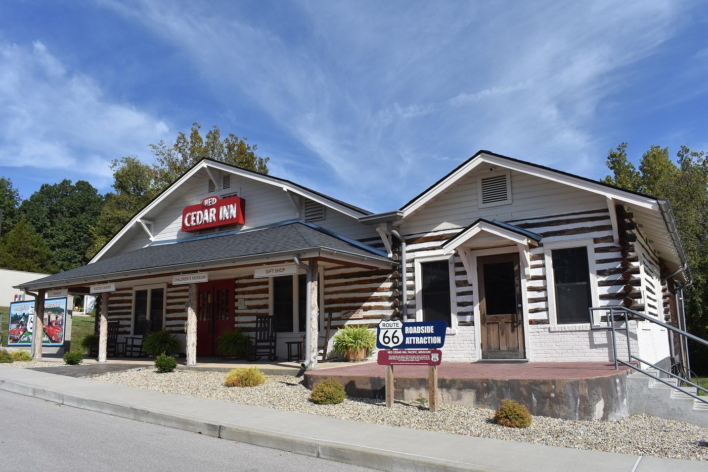 A restored log building with white chinking, a red Red Cedar Inn sign, a covered entry porch and a Route 66 Museum sign on the sidewalk under a blue sky