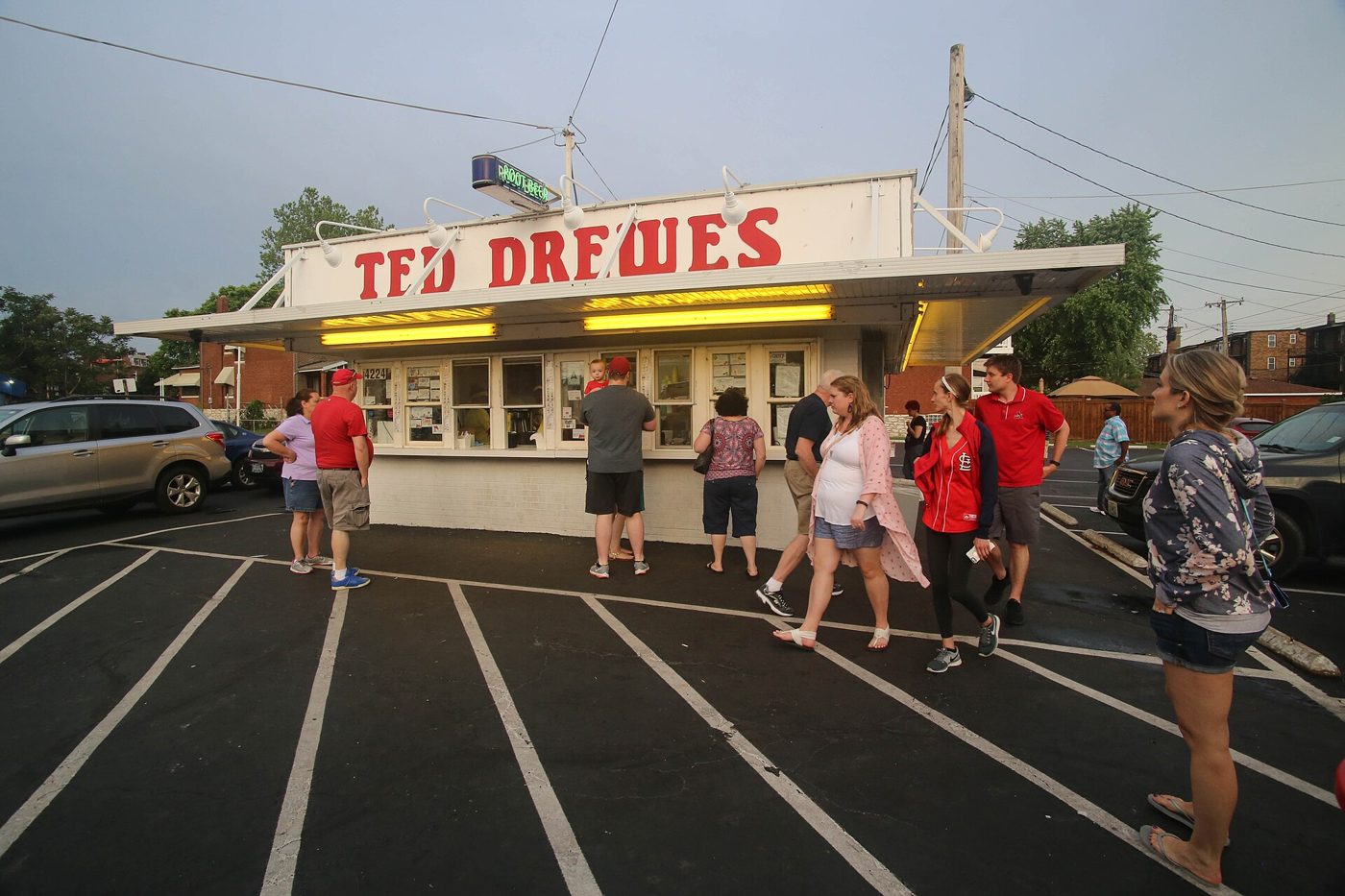 A white single-story custard stand with a long row of service windows and a yellow Ted Drewes sign, with customers standing in line in the parking lot