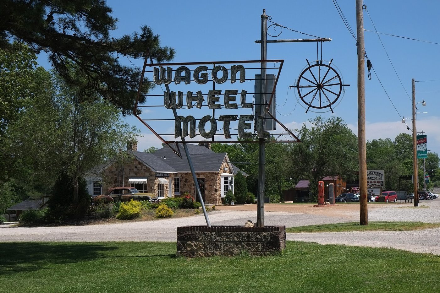 A tall roadside sign with Wagon Wheel Motel in neon script and a metal wagon wheel, with stone cottages and trees behind it