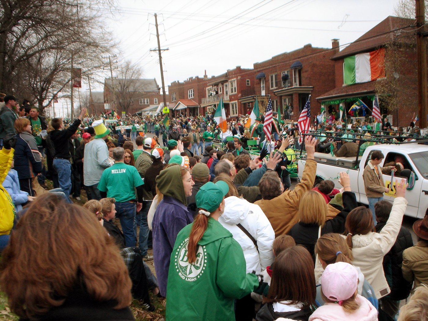 Crowd in green crammed along a brick residential street, Irish and American flags flying from a porch, a pickup pulling a float past waving spectators