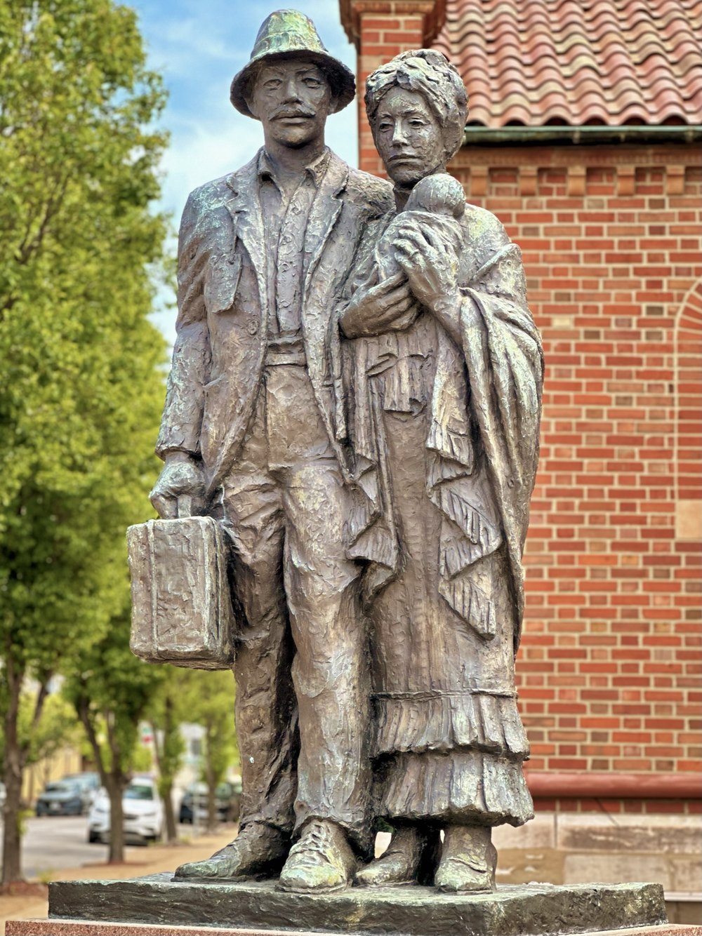 Bronze sculpture of a man in a cap holding a suitcase beside a woman in a shawl holding an infant, standing in front of a brick church wall