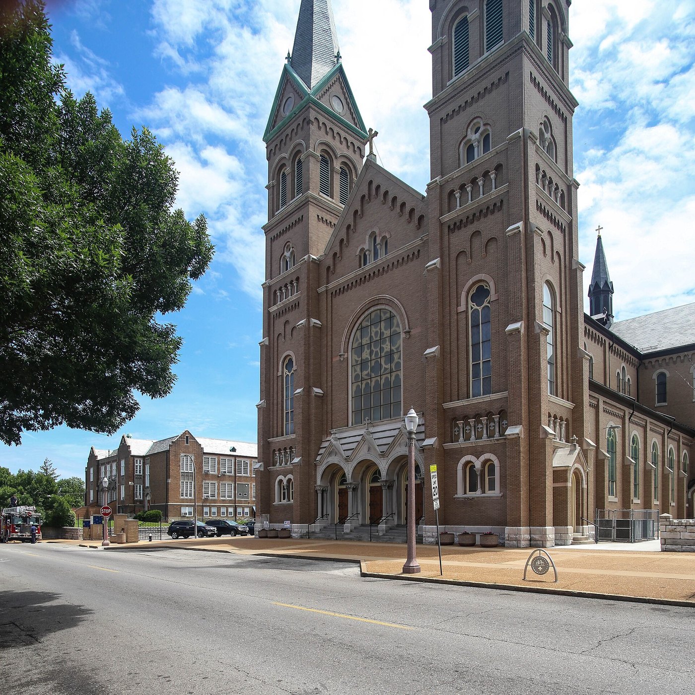 Large red-brick Romanesque church with twin pointed spires rising above a tree-lined South St. Louis street
