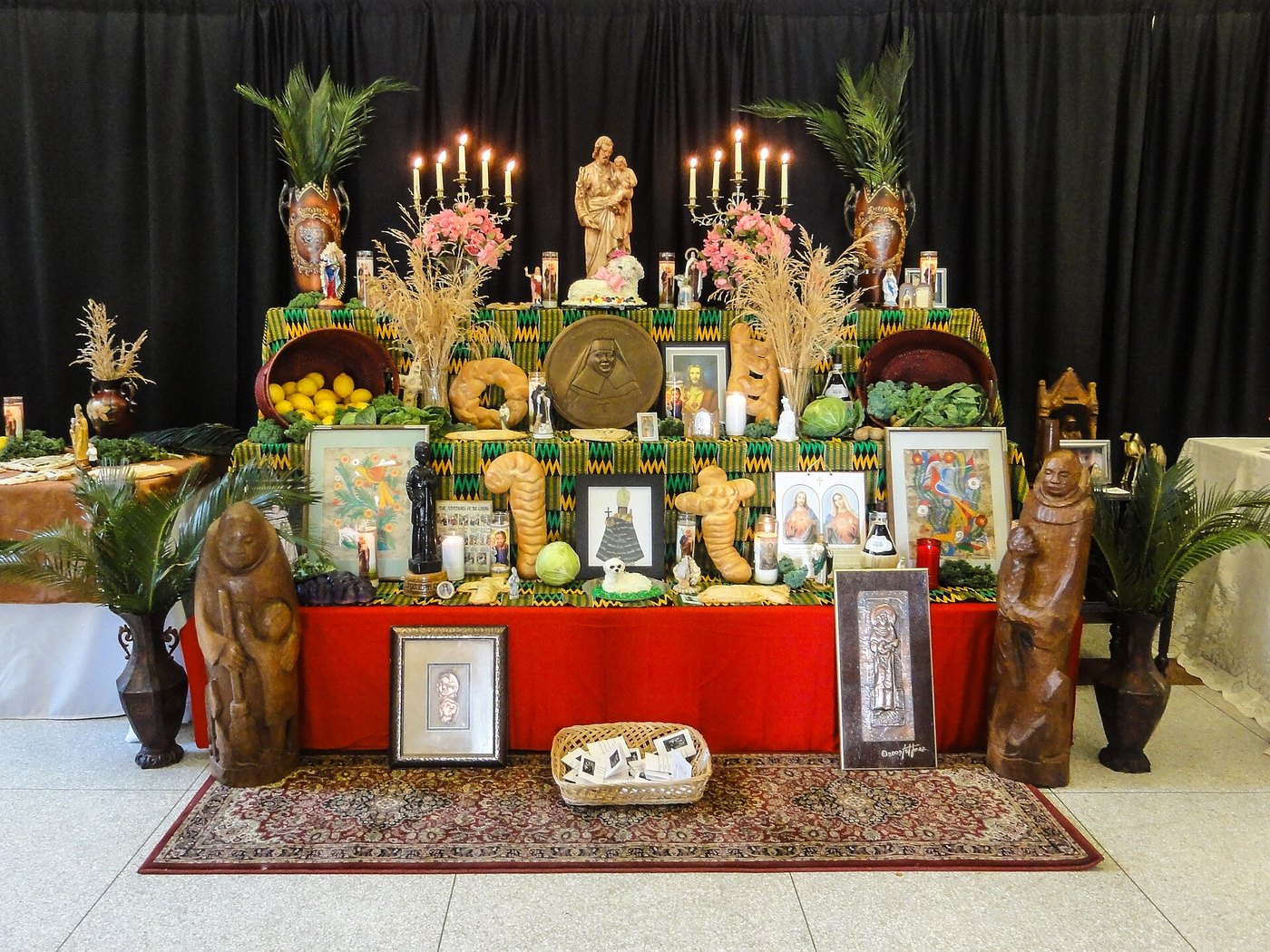 Three-tiered altar draped in red and green, crowded with shaped breads, cakes, fruit, candles and framed holy pictures, a statue of St. Joseph at the top